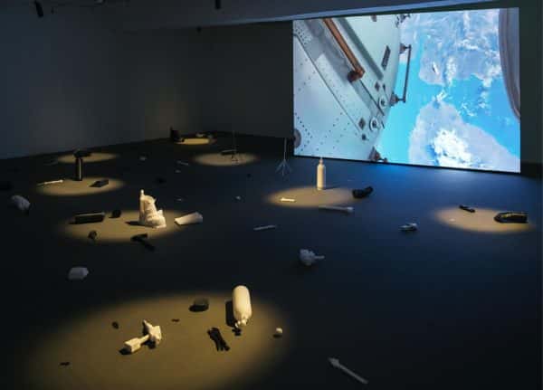 Drifiting - Objects scattered across a dark floor are spotlit from above, in the background a large installed screen shows footage from a man-made craft orbiting earth. The objects look to be recreations of astronauts equipment and debris from space wreckage