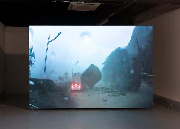 Falling Rock installation - A large LCD screen installed in an empty room, shows footage through a car windscreen, obscured by dust and raindrops, shows a vehicle with brake lights on, driving round an enormous boulder in the middle of the road. The environment is dusty and feels like a disaster zone