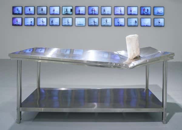 Table Series - A sturdy metal table crumpled on one side, supports a section of marble pillar. In the background, two rows of LCD screens show photographs featuring the table, some before and some after its destruction