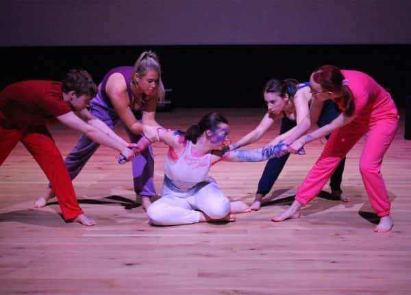  - A group of four pull the ams of an actor kneeling on the floor, the performer has paint on their arms and face