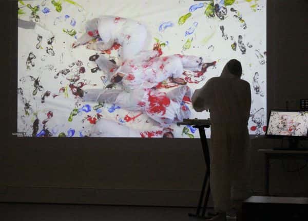  - Someone in a white overalls looking at a mock crime scene photo on a projected screen, where more people in overalls lay dramatically strewn across a white sheet. The sheet and the people in the image are all covered in footsteps of colourful paint