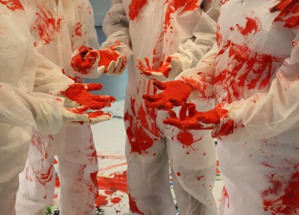  - A group of people wearing white coveralls and white gloves stand in a circle with their hands in the middle, palms facing up, covered in red