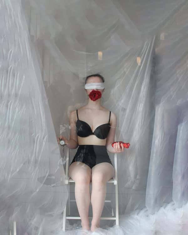  - An actor in modest black underwear sits on a white chair in a room with white backdrop and white fabric on the floor, holding an open pocket watch in one hand, and what looks like a heart in the other. They are blindfolded with white fabric and have the bloom of a large red rose covering the lower half of their face, as though planted in their mouth. The scene is behind a layer of something transparent like glass, and you can see the reflection of people in the background of the room watching