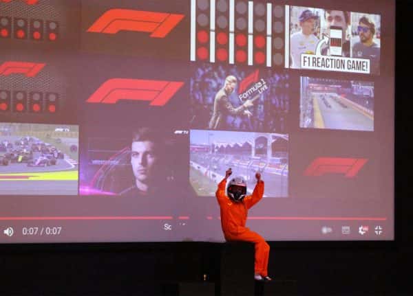  - Someone with orange coveralls and a racing helmet sits on a dark stage, backdropped with a huge screen showing a grid with various scenes from a formula one race