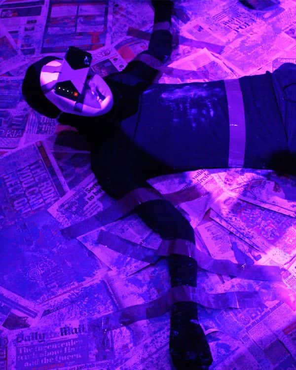  - A figure under bright purple light, is taped to a surface of tabloid newspapers wearing an expressionless mask which has a mobile phone fixed over the eyes