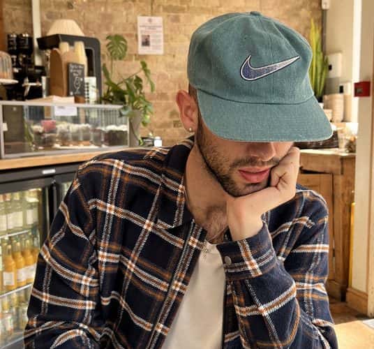  BA Fashion Communication and Promotion student Joe Corlito is sat at a wooden table in a modern cafe, working on his laptop. He is looking down at his screen, wearing a sage green baseball cap that covers half of his face