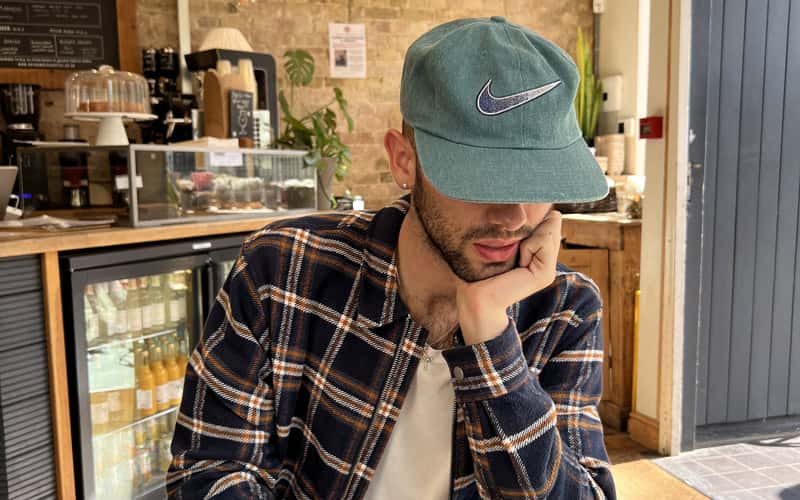 BA Fashion Communication and Promotion student Joe Corlito is sat at a wooden table in a modern cafe, working on his laptop. He is looking down at his screen, wearing a sage green baseball cap that covers half of his face