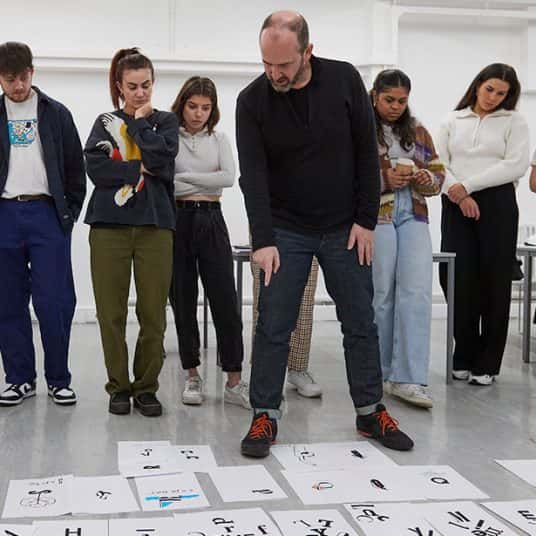  Image showing person leading workshop talking through student work on floor with students in background