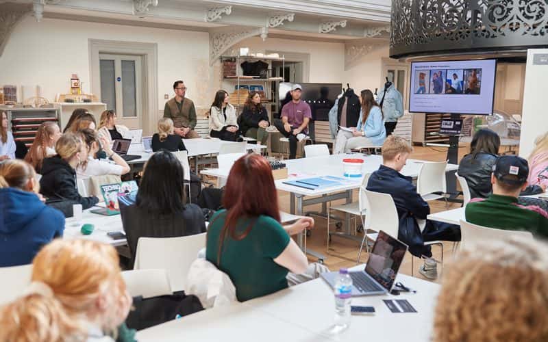 A photograph of the interior design studio at Boardman House, Norwich. A TV screen is placed near the staircase in the centre of the room, and has a presentation of fashion images from clothing brand Blakeley. Sat next to the TV are NUA staff and Blakeley employees. They are presenting a project brief to a class of Interior Design and Fashion Marketing and Business students