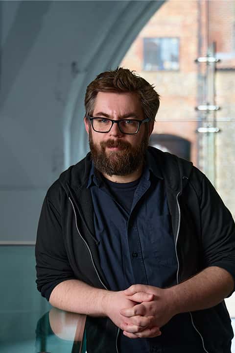Games Art and Design Lecturer Ian Griffiths with short brown hair, thick beard, and glasses, standing in a Norwich University of the Arts computer lab