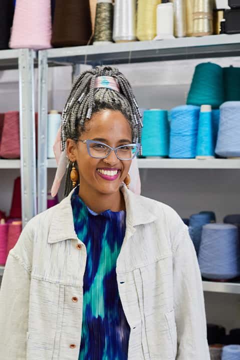 Senior Lecturer Odette Steele stands for a photo in front of shelves which are full of colourful reels of thread and yarn. Odette wears pastel cat-eye frame glasses, and a light blue denim jacket over dark blue tie-die pattern top