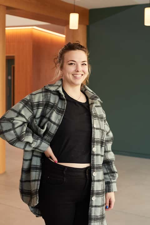 Games Art and Design lecturer Michelle Marham stands in the Duke Street Riverside lobby. She wears a neutral green and grey checkered flannel shirt over a black vest and black jeans