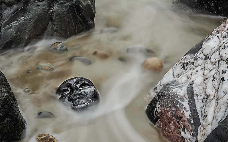 Photography by Paul Mills shows an eerie mask with blank expression, laying beneath shallow waves on a sandy shore. There are some pebbles between large smooth marbled boulders, and the small waves look like smoke where a long exposure timelapse has blurred the movement around the pebbles.