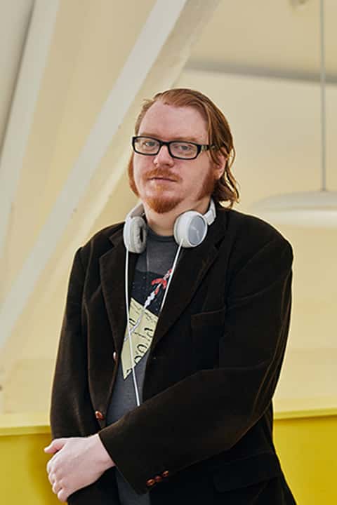 Portrait image of Dr Dean Bowman in the media lab at Norwich University of the Arts. Dean is leaning against the wall, wearing a dark cotton cord jacket over a graphic tee