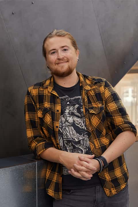 Games Development Lecturer George Britton leans against the sheet-metal handrail of the Guntons Building staircase. George is smiling, and is wearing a dark yellow and black check over-shirt over a black graphic tee