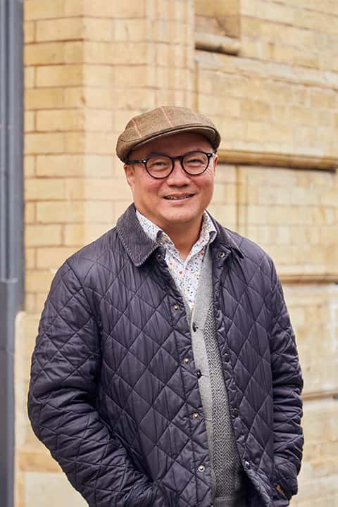Architecture Lecturer William Chen stands in front of the St Georges building at Norwich University of the Arts. William wears a flat peaked cap and navy quilted jacket