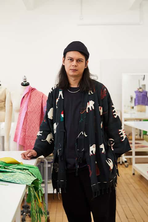 Fashion Course Leader Jasper Chadprajong-Smith stands in a Fashion workshop with some colourful fabrics. Jasper has shoulder length black hair, and wears a black beanie, loose black t-shirt, black trousers, a silver chain, and an open black cardigan patterned with ignorant style animal illustrations