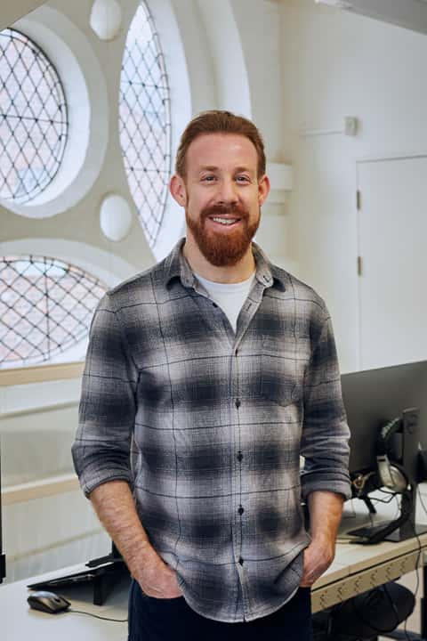 Staff profile image of Dan Kelby. Dan wears a plaid shirt over a white top, and is stood in front of the windows in the Media Lab