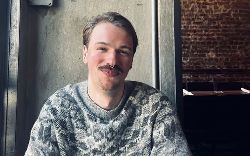 Photo of Animation alumni August Abrahamsson. August is smiling at the camera, wearing a grey jumper and sitting with his arms folded. There is a grey wall behind him and a brick wall can be seen through a door to his left.