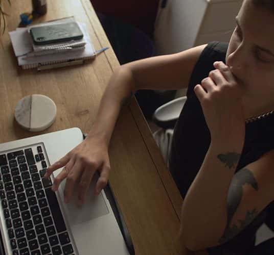  Still from film 'Is There Anybody Out There?' dir. Ella Glendining. Top down view of Ella Glendining, who sits at a desk with one hand on the trackpad of a laptop. She is wearing a black vest and has tattoos on one arm.