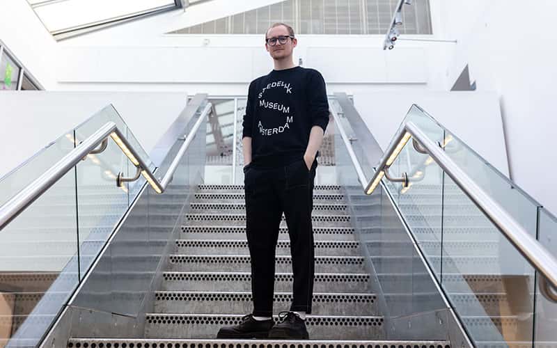A full body portrait of Jeremy Downes standing on a flight of stairs, looking straight at the camera and dressed in all black.