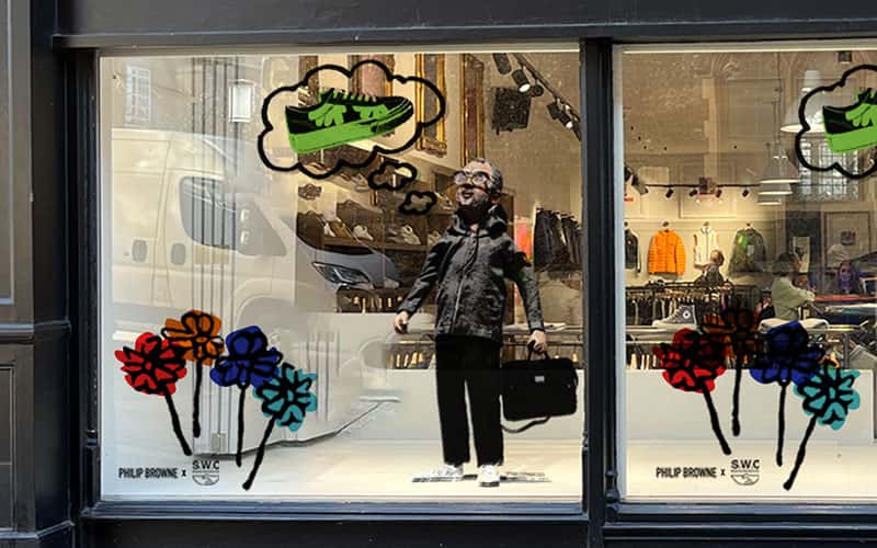 A photo of Philip Brownes shop window with an illustration of Philip Brown thinking about Stepney Workers club trainers in a thought bubble. The illustration of Philip is also holding a briefcase and standing next to four large flowers coloured red, orange, purple and blue.
