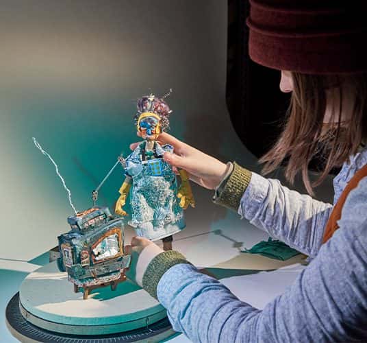  A close-up photograph of an animation student working in a studio environment. The work is a small stop motion character and tv.