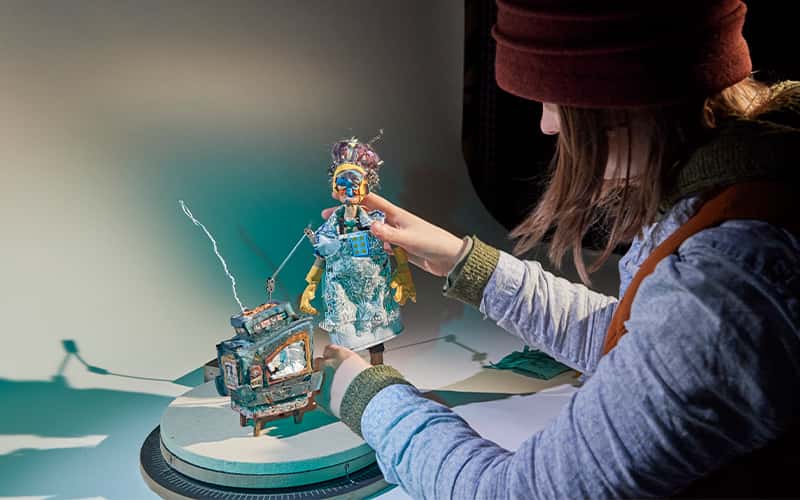 A close-up photograph of an animation student working in a studio environment. The work is a small stop motion character and tv.
