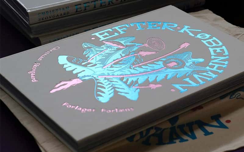A hardback book laying flat. The book is named Efter København which is in large blue foil writing, along with a blue and pink foil plant illustration.