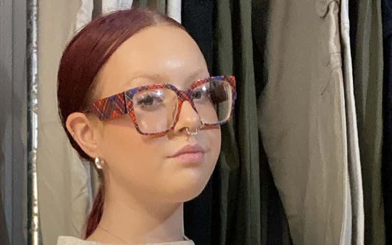Portrait of Fashion and Communication student Yasmin Welch, with red dyed hair tied back in a pony tail, and wearing thick rimmed cat eye glasses in a tartan pattern