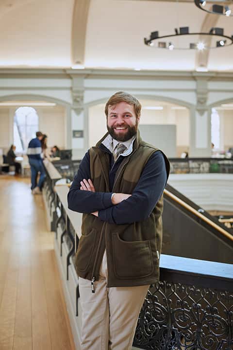 Architecture Lecturer Matt Sawyer stands smiling for staff portrait wearing a dark green gilet over a shirt, tie, and jumper