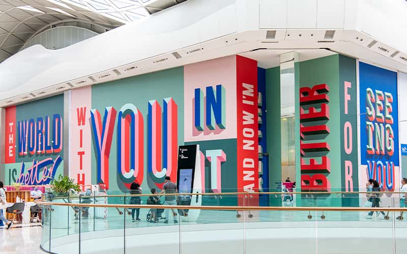 A photo of Rachel Joy Prices artwork in London Westfield shopping centre. The brightly coloured lettering has been printed onto empty shop units.