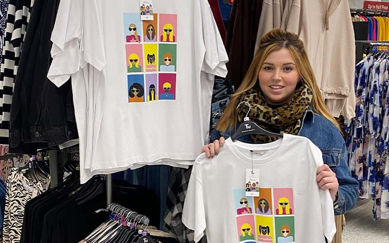 A photo of Evie Thomas in a Sainsbury's store holding a t-shirt with her design on it.